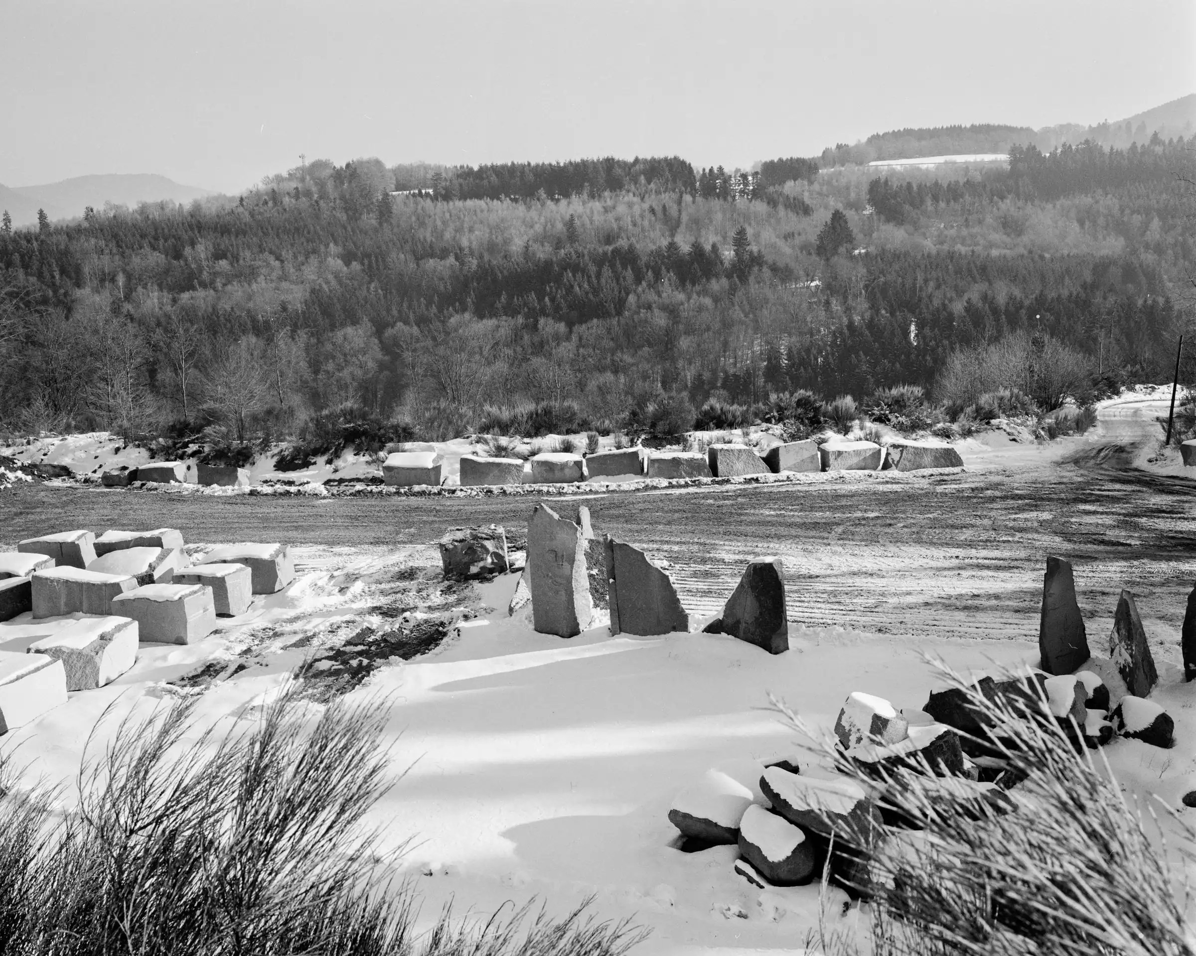 Black-and-white snowy quarry yard with stacked stone blocks and forested hills.