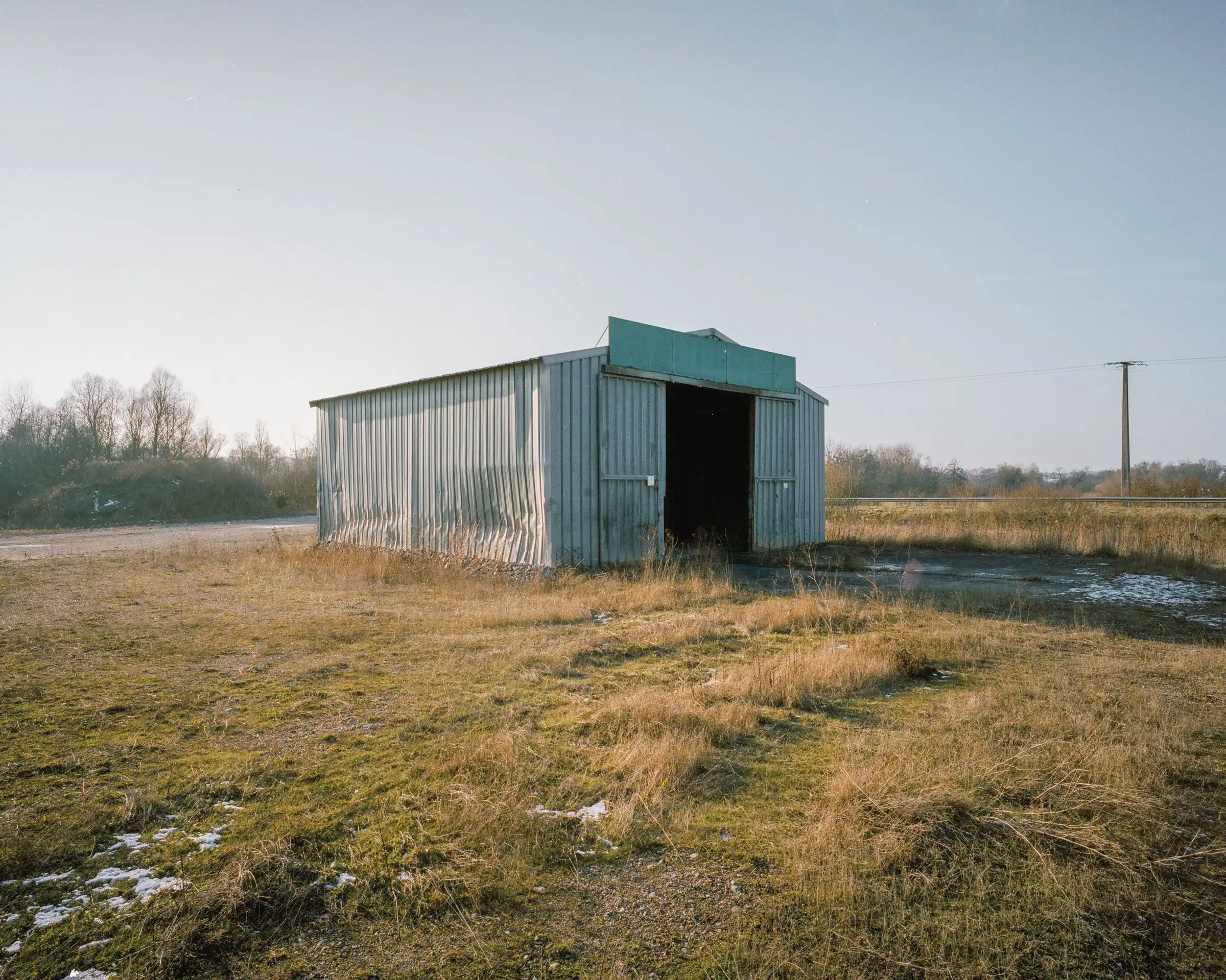 Corrugated metal shed with open doorway in a dry grassy field and pale sky.
