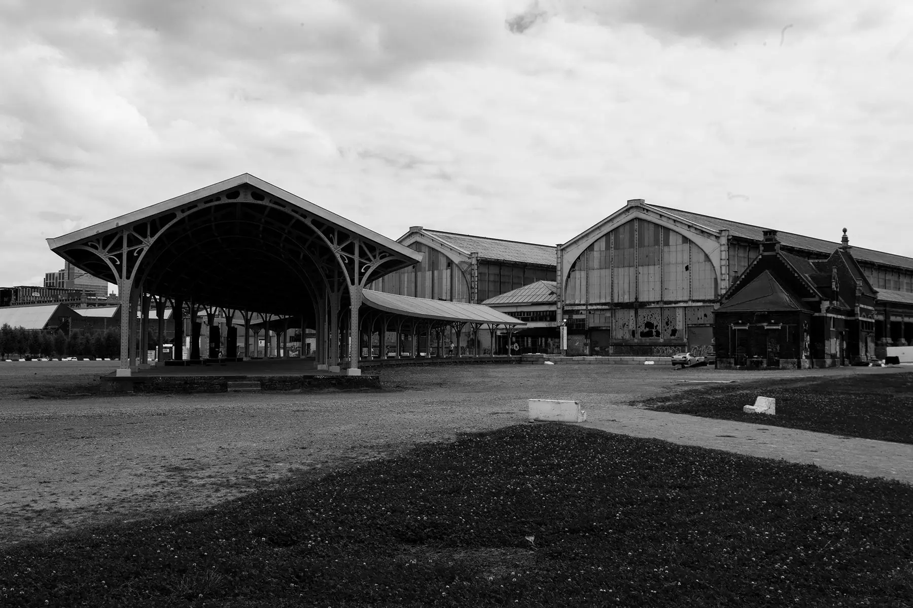 Black-and-white view of an open metal canopy and warehouse buildings under cloudy sky.