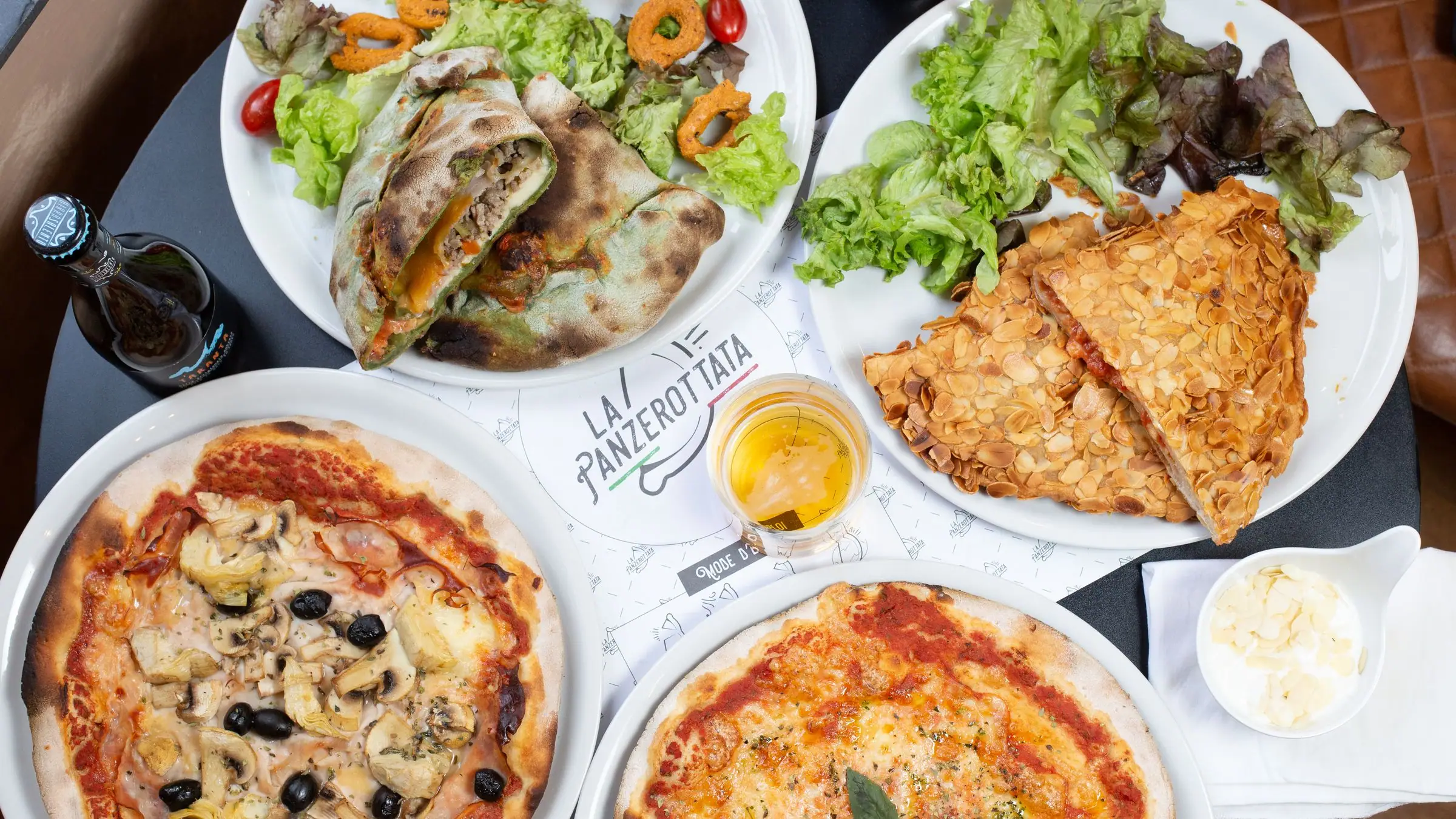 Top-down spread of panzerotti, two pizzas, salad, beer bottle and glass on a dark table.