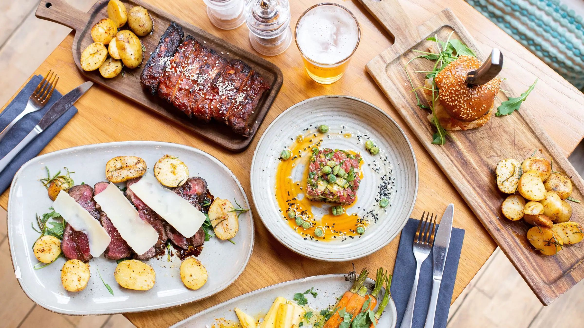 Flat lay of ribs, burger, steak, tartare and beer on a light wooden table.