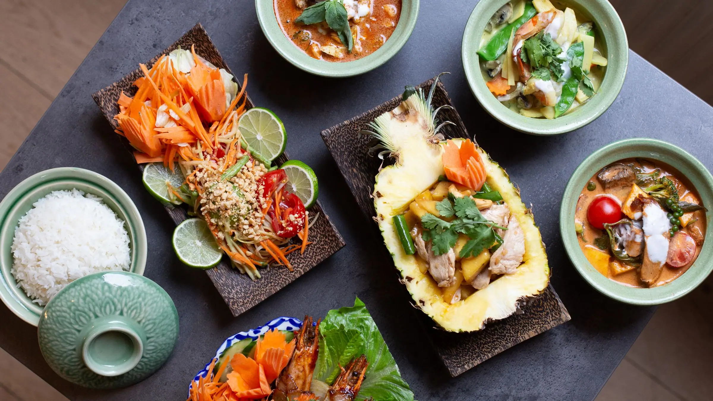 Flat lay of Thai curries, papaya salad, pineapple boat and grilled prawns.
