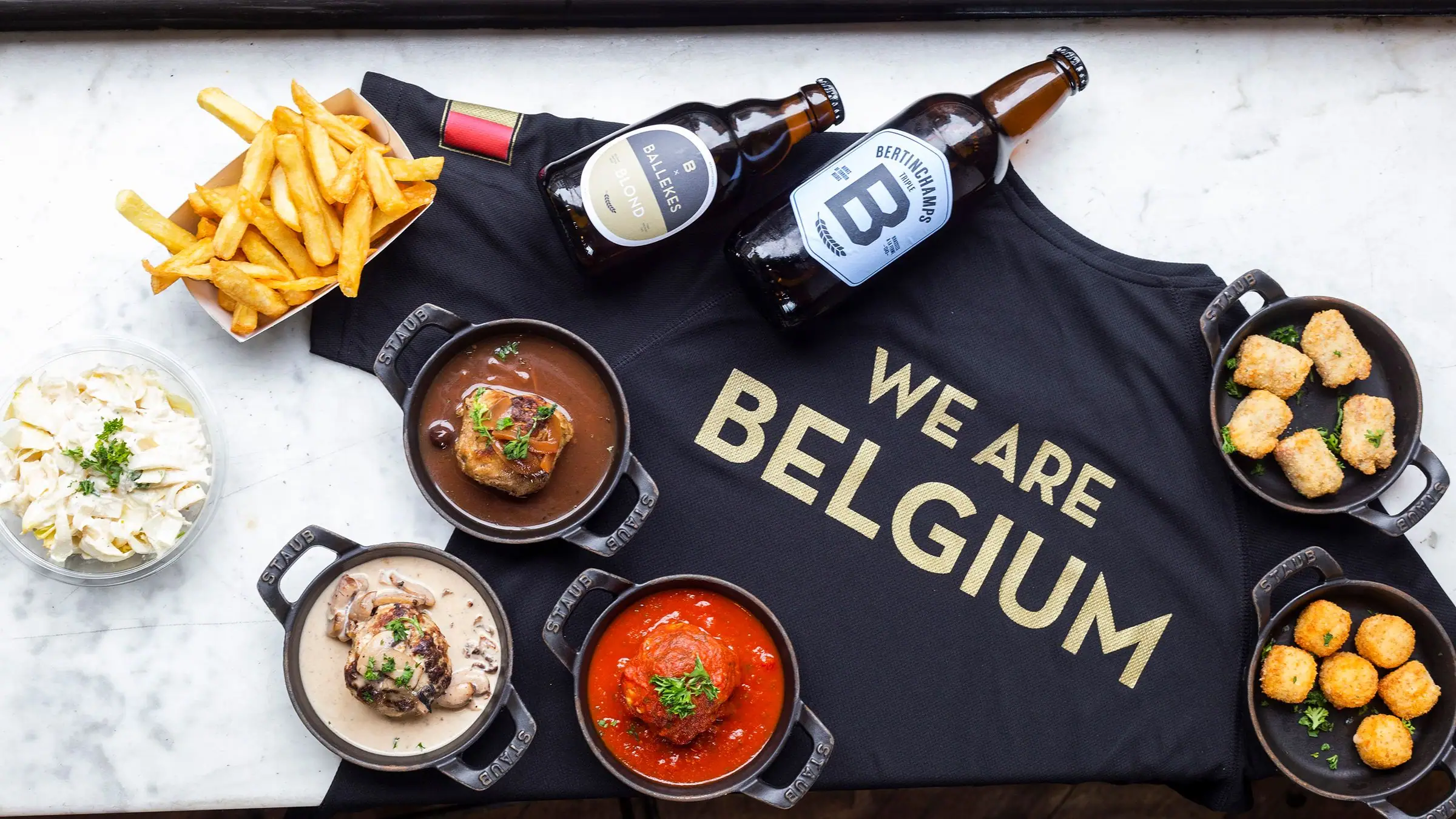 Flat lay of Belgian meatballs, fries and beers around a “WE ARE BELGIUM” shirt.