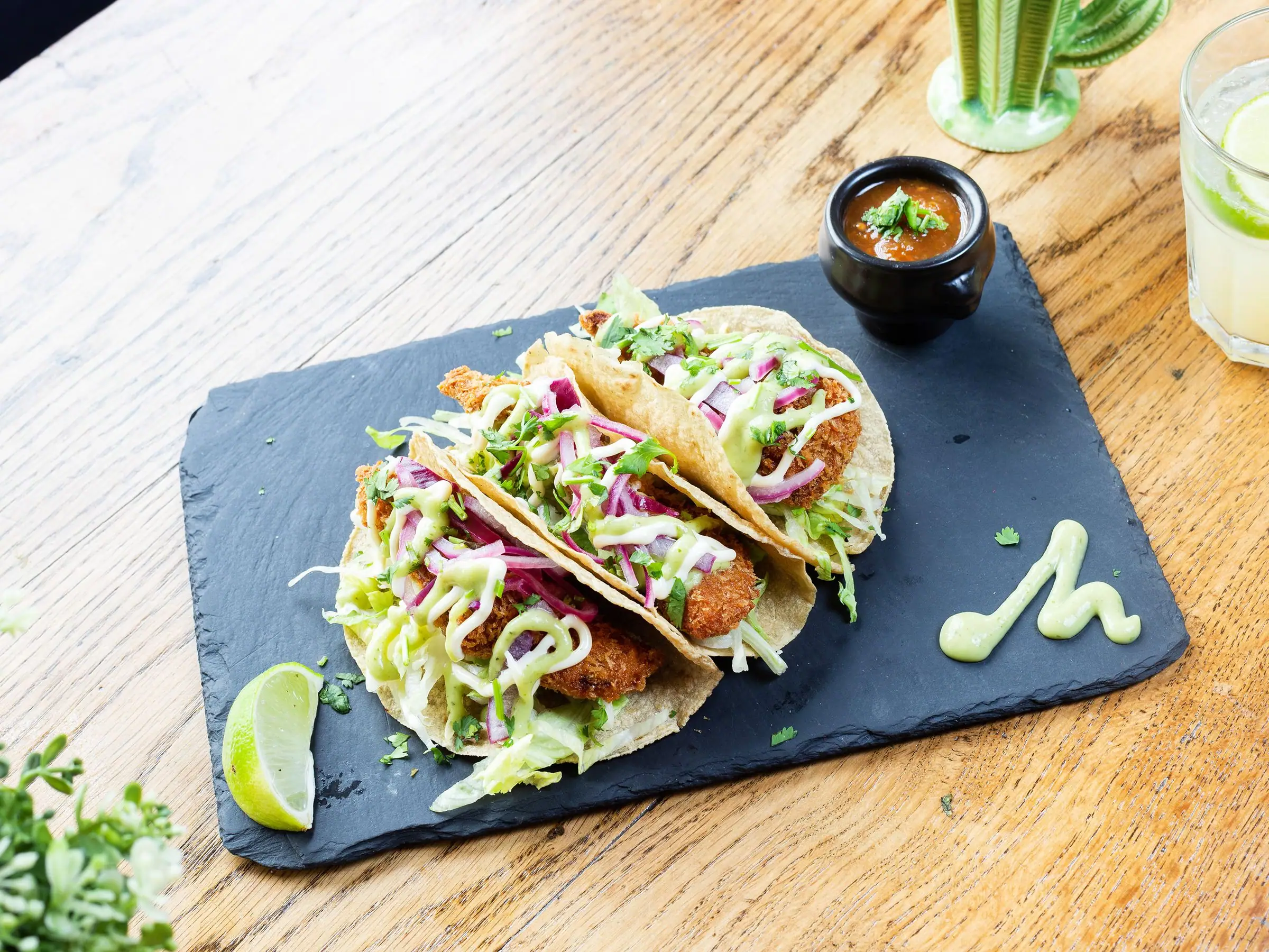 Three fried-chicken tacos on slate with lime, salsa and green sauce drizzle.