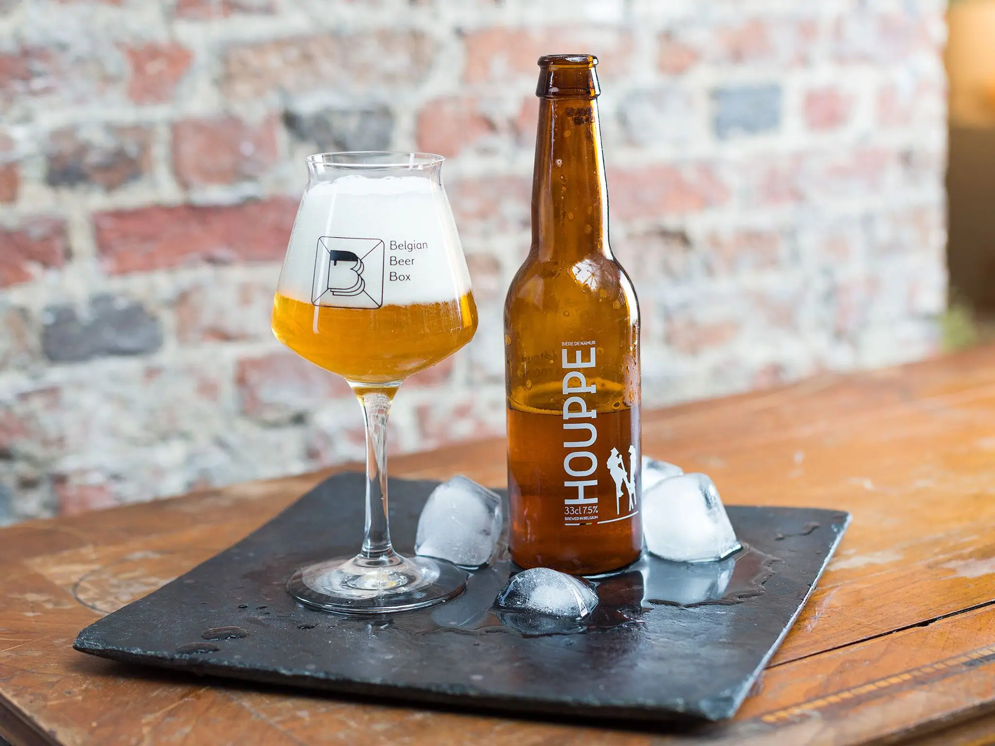Houppe beer bottle and filled glass on slate with ice cubes, brick wall behind.