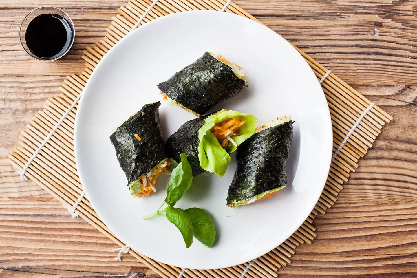 Four nori rolls with vegetables on a white plate beside a small sauce bowl.