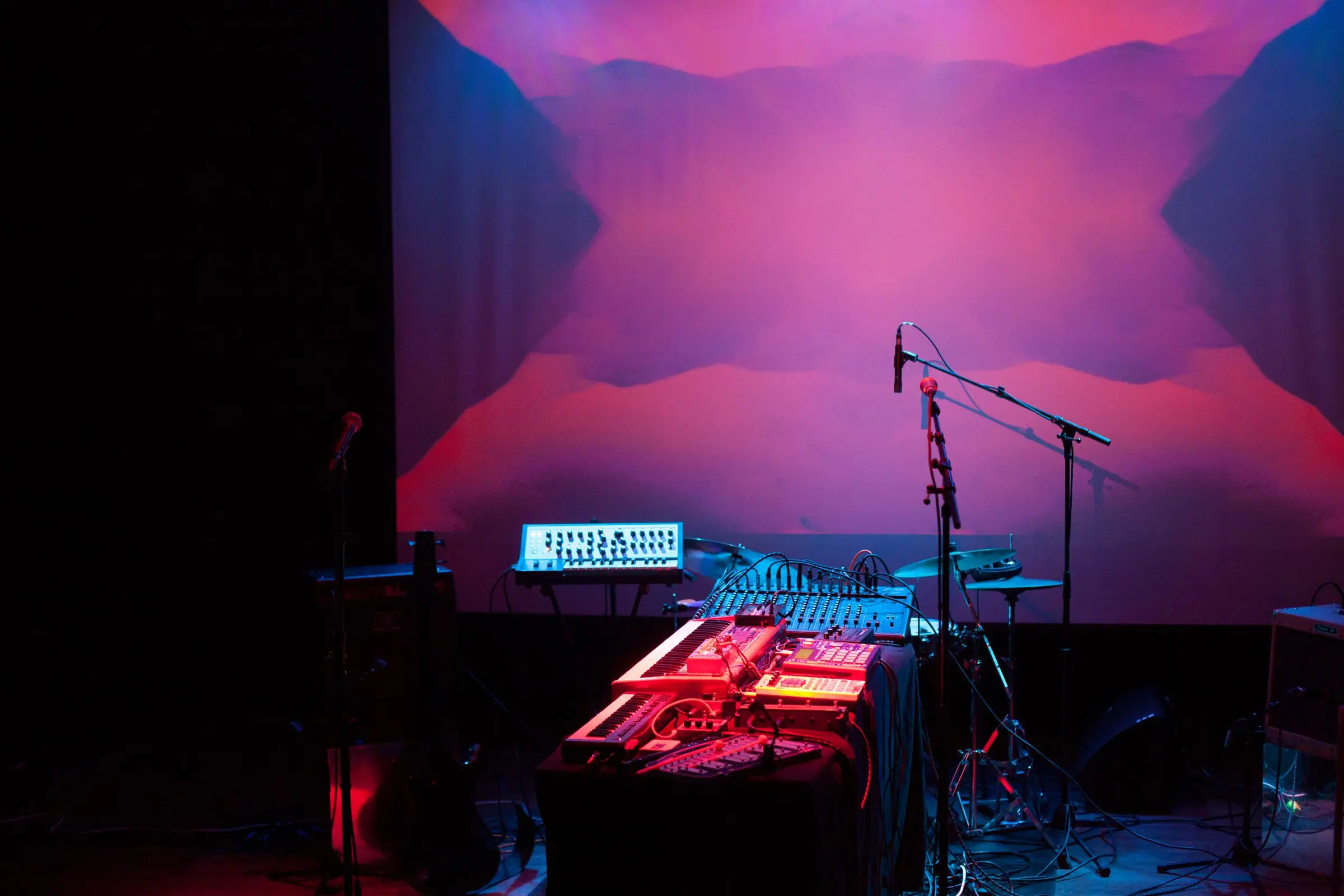 Dark stage with synths and mixer under red and blue lights, abstract screen behind.