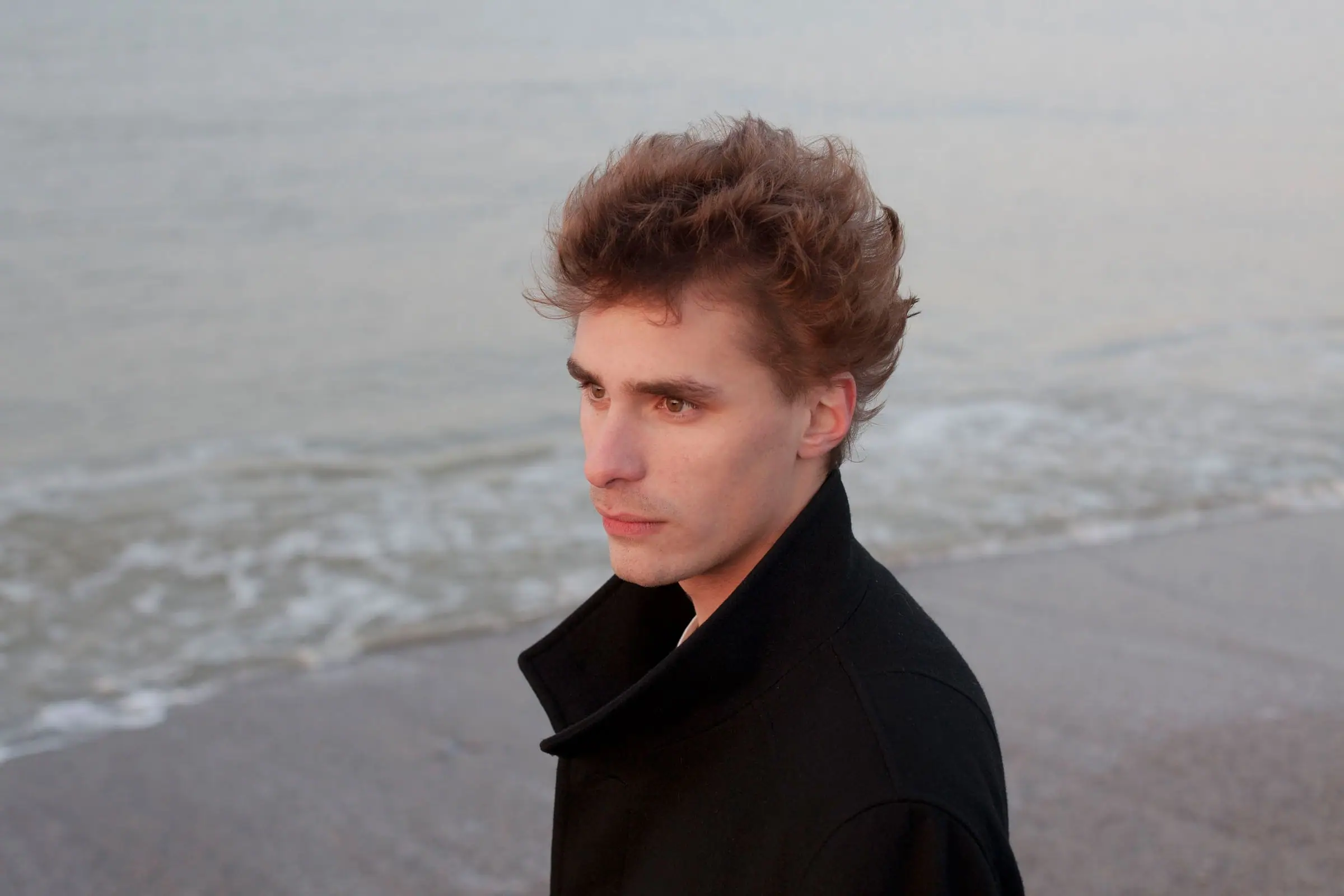 Portrait of a young man in a dark coat looking aside, blurred beach behind.