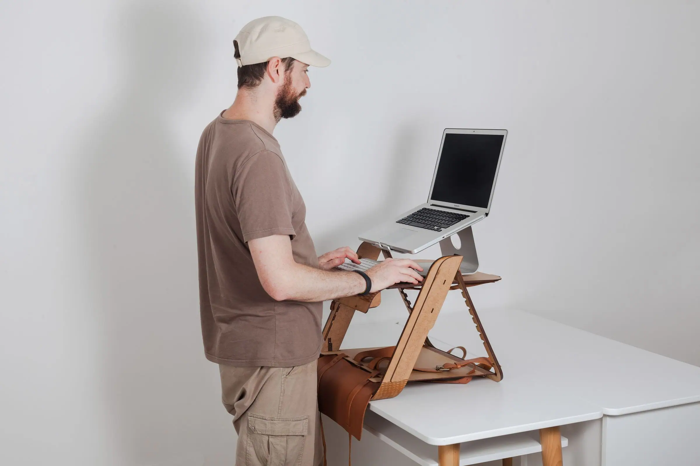 Bearded man in a cap typing at a plywood standing desk with a laptop.