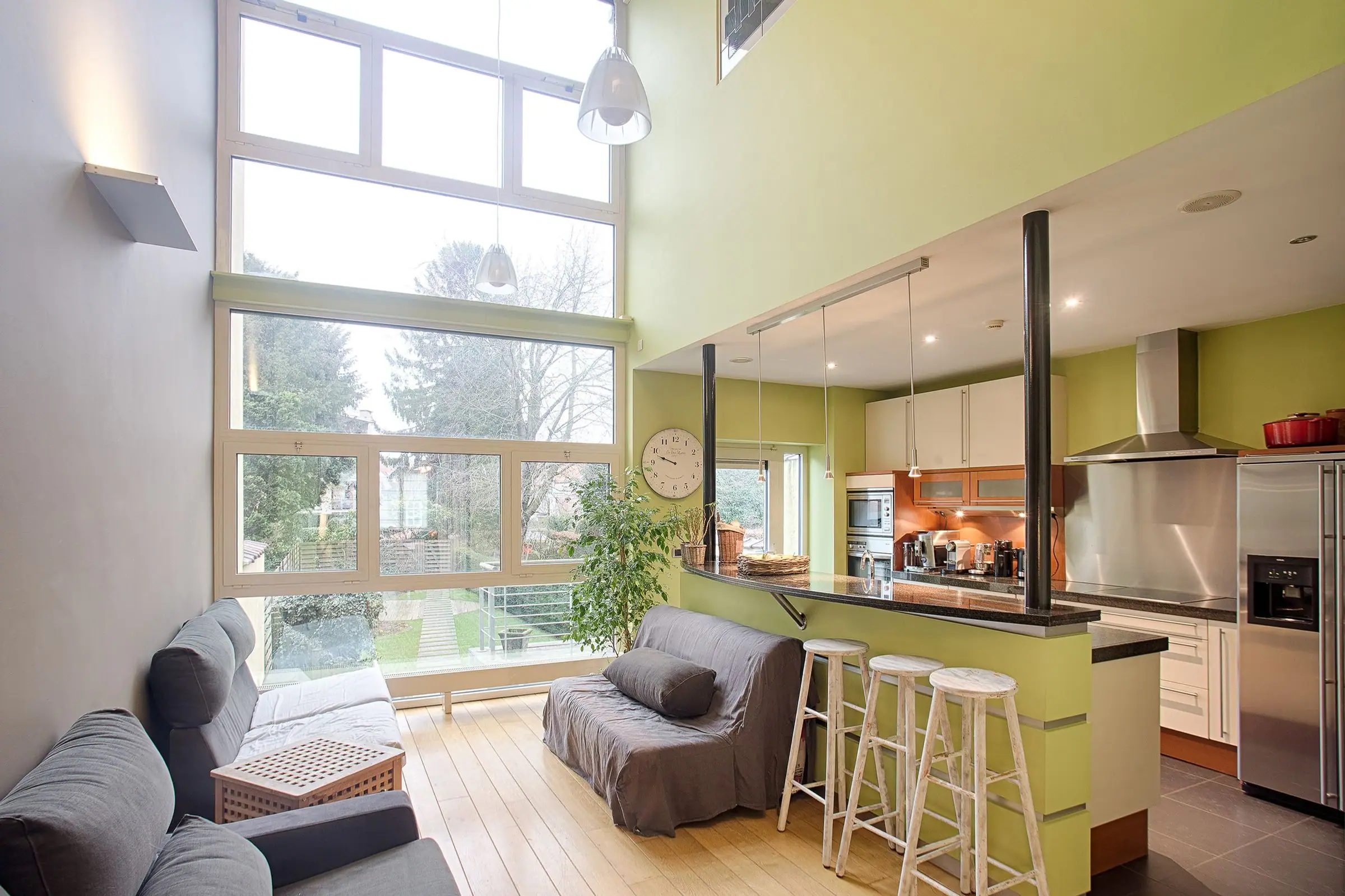Open-plan living room and kitchen with high windows, green walls and bar stools.