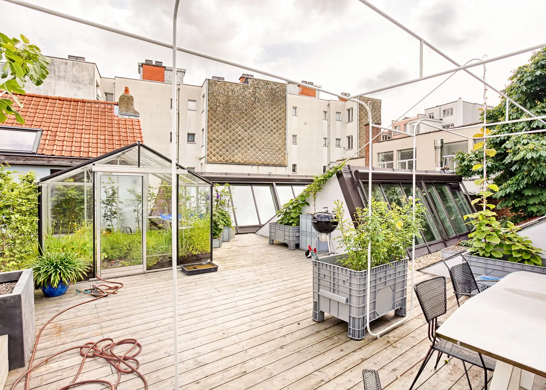 Urban rooftop terrace with glass greenhouse, planters, grill and outdoor seating.