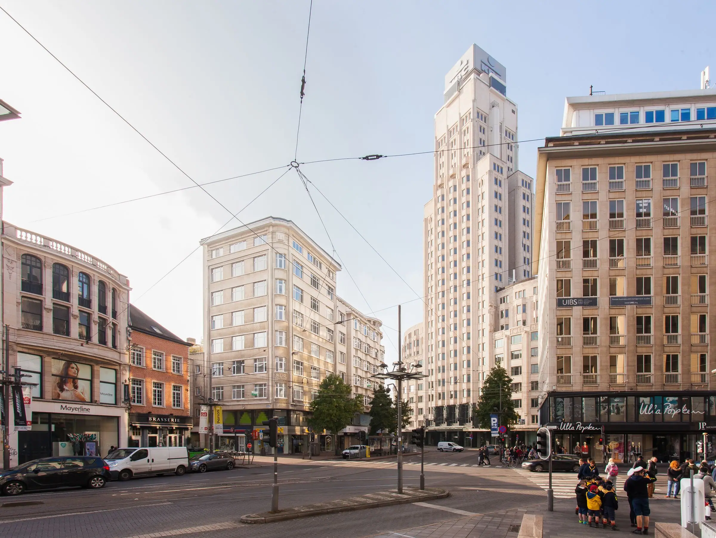 City intersection with tall white skyscraper, shopfronts and overhead tram lines.