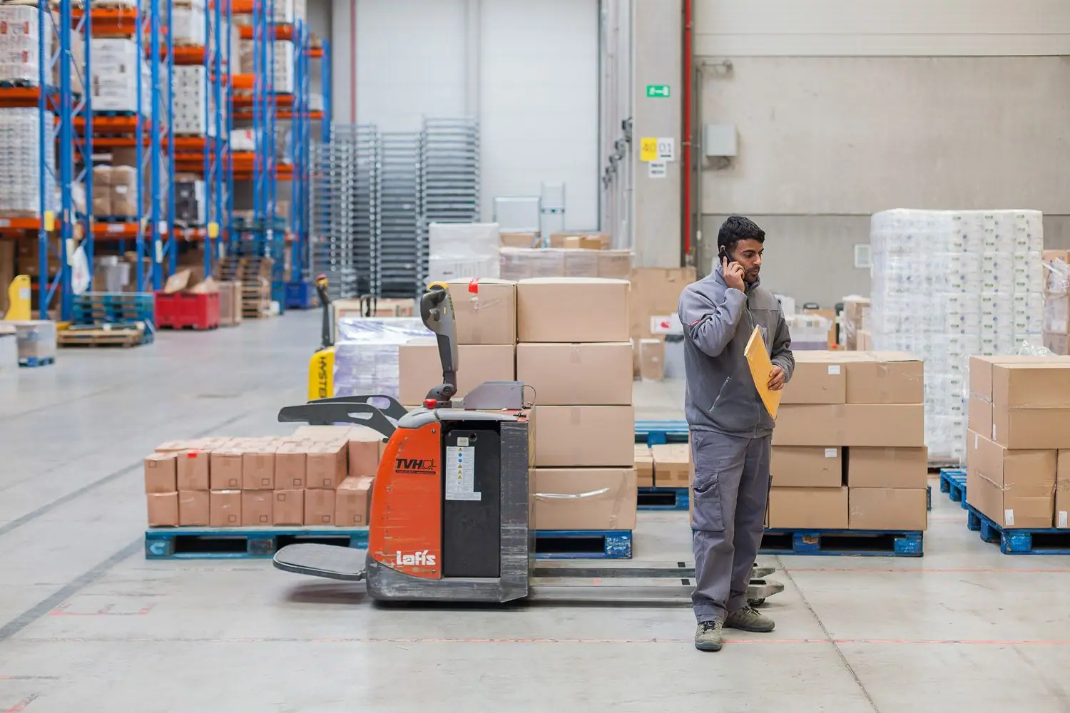 Warehouse worker on phone holding a yellow folder beside an orange pallet jack.