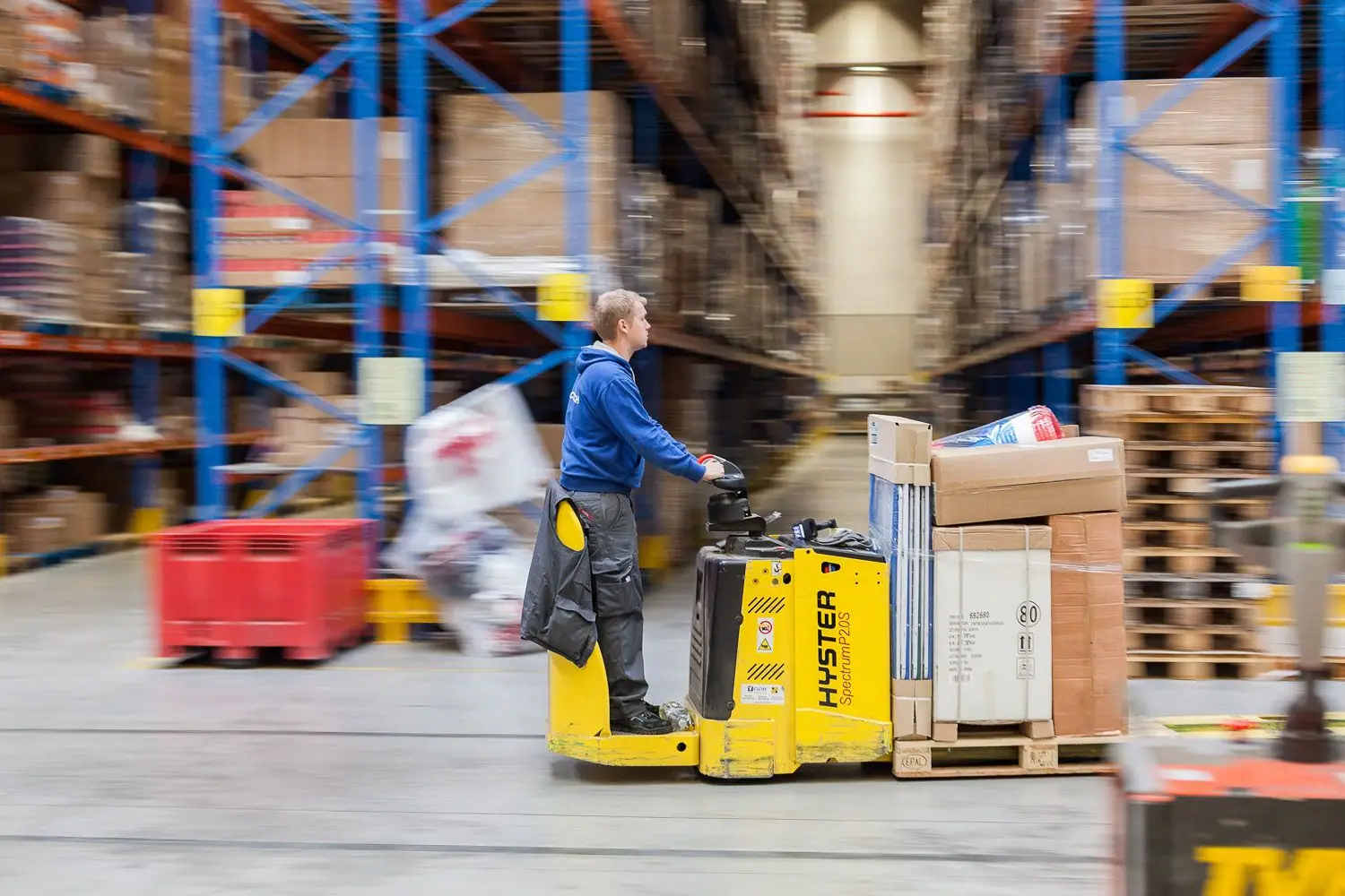 Worker riding a yellow pallet jack with boxes, motion-blurred warehouse racks.