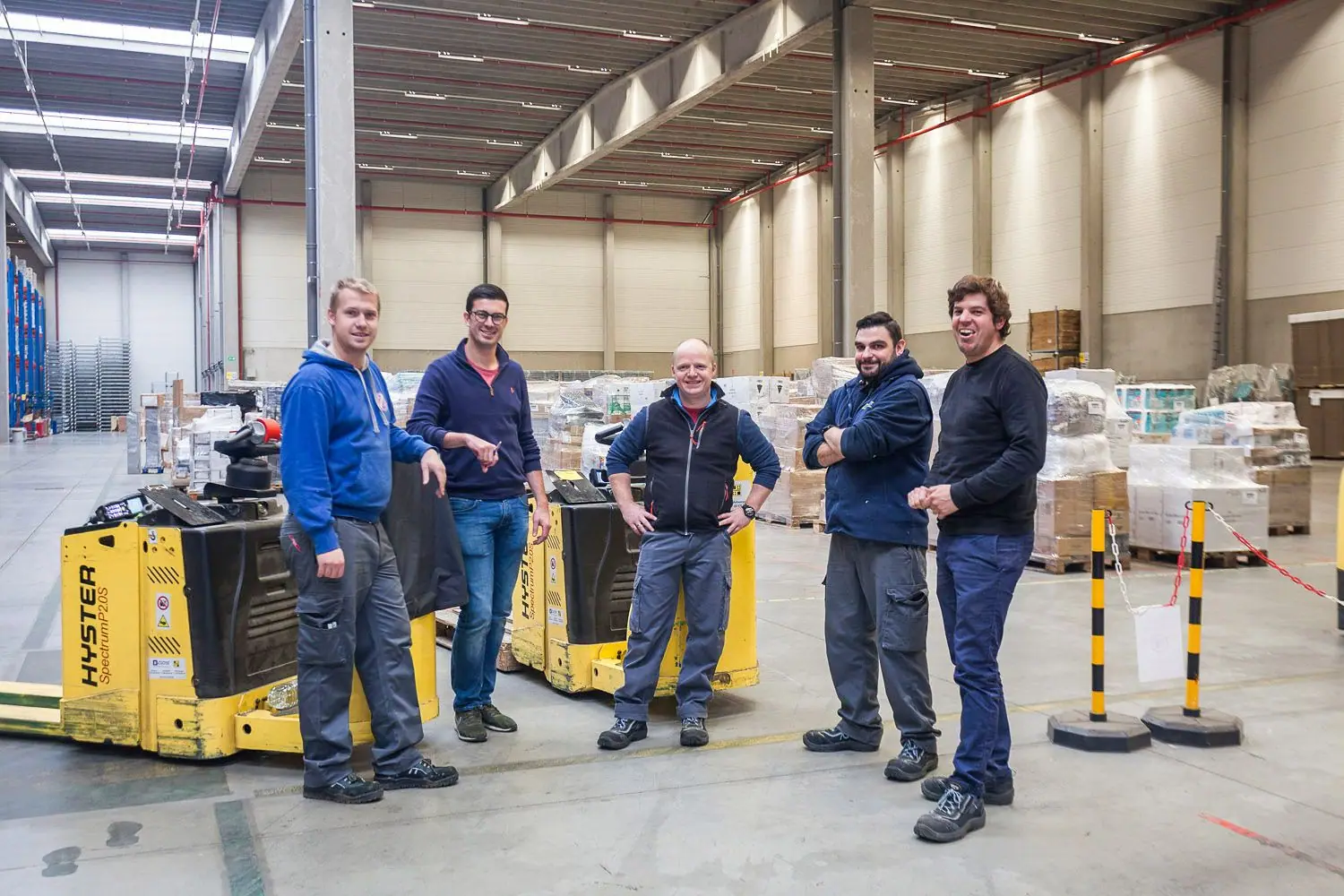 Five warehouse workers standing together near a yellow pallet jack and stacked pallets.